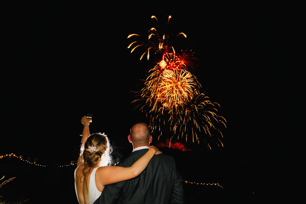 the bride lifts her glass whilst hugging her groom whilst fireworks go off in the background