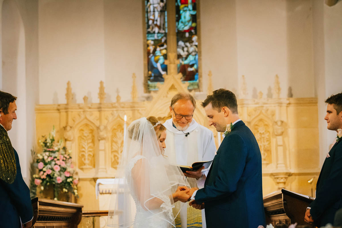 the bride puts the ring on the grooms finger, the vicar smiles behind