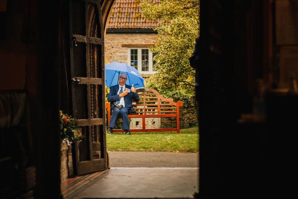 a wedding guest gives the photographer the thumbs up while he waits in the rain in the churchyard outside the ceremony