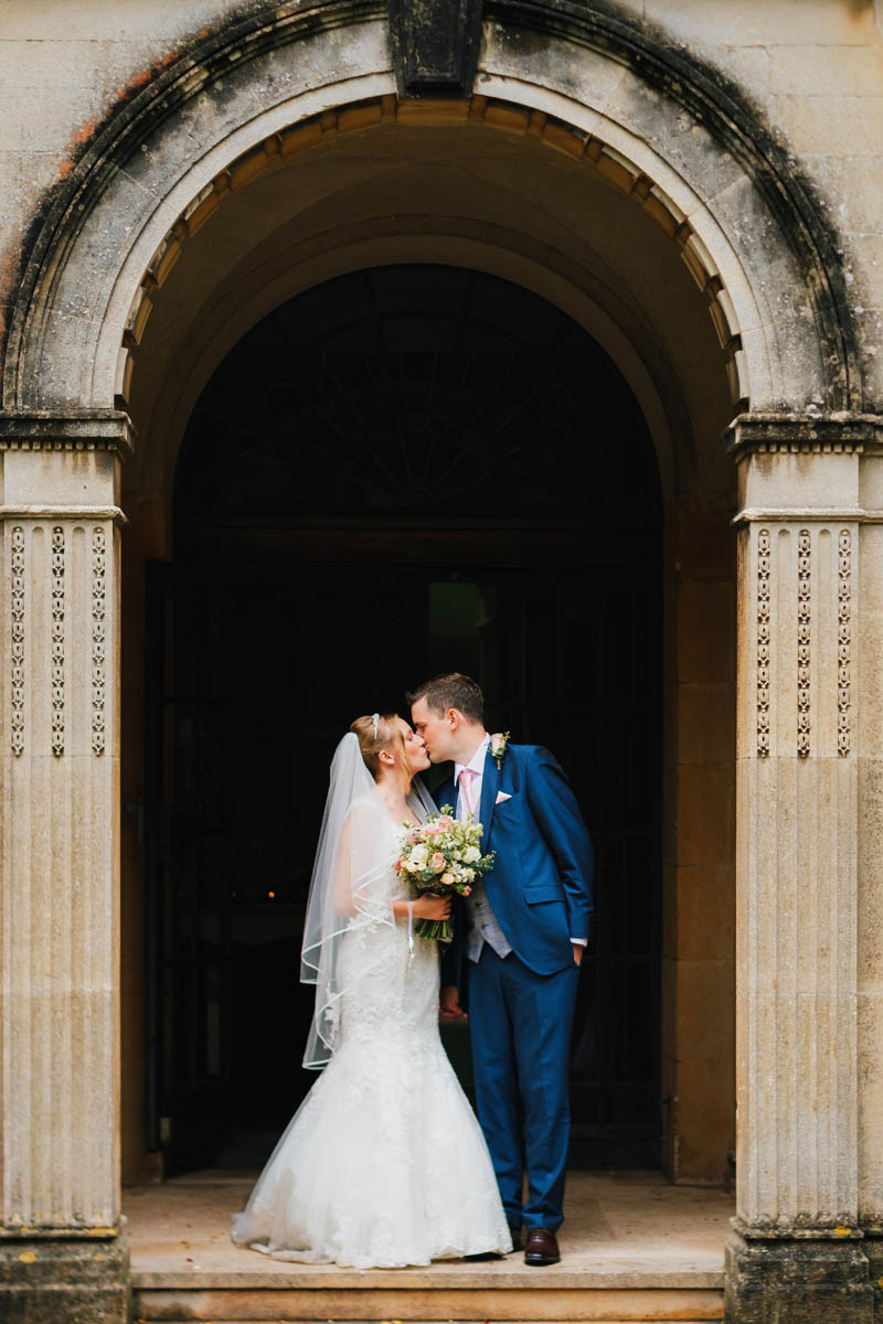 bride and groom kiss in an archway of the building, the bride holds her bouquet