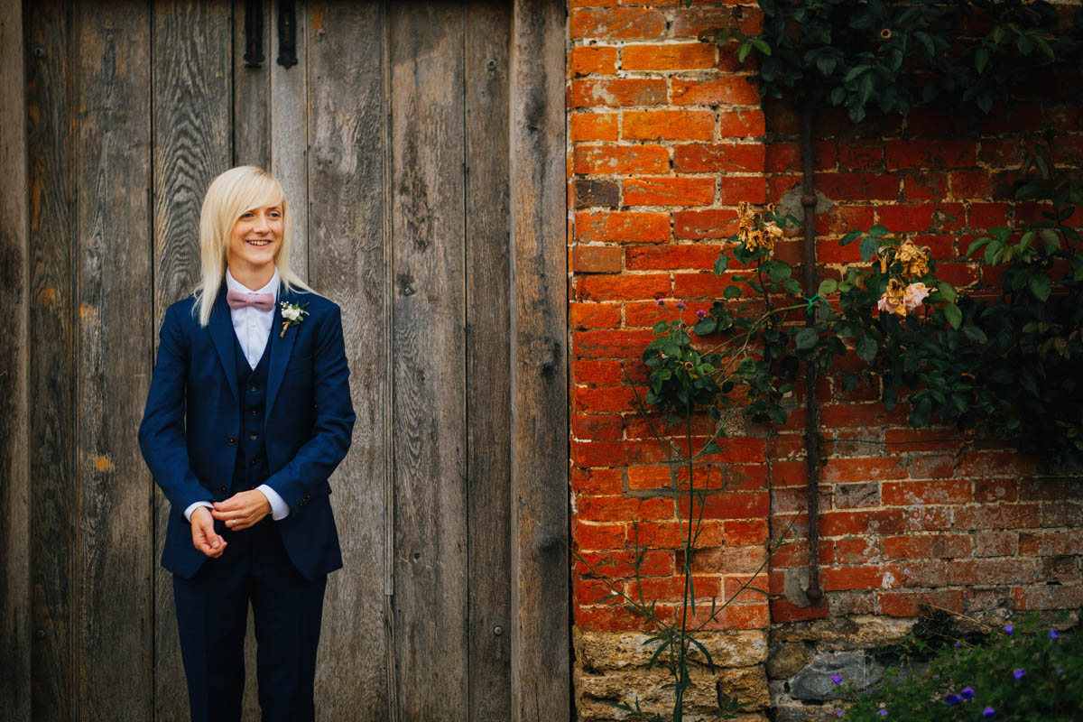 the bride in her suit looking off to one side, there is a redbrick wall and rustic wooden door behind her