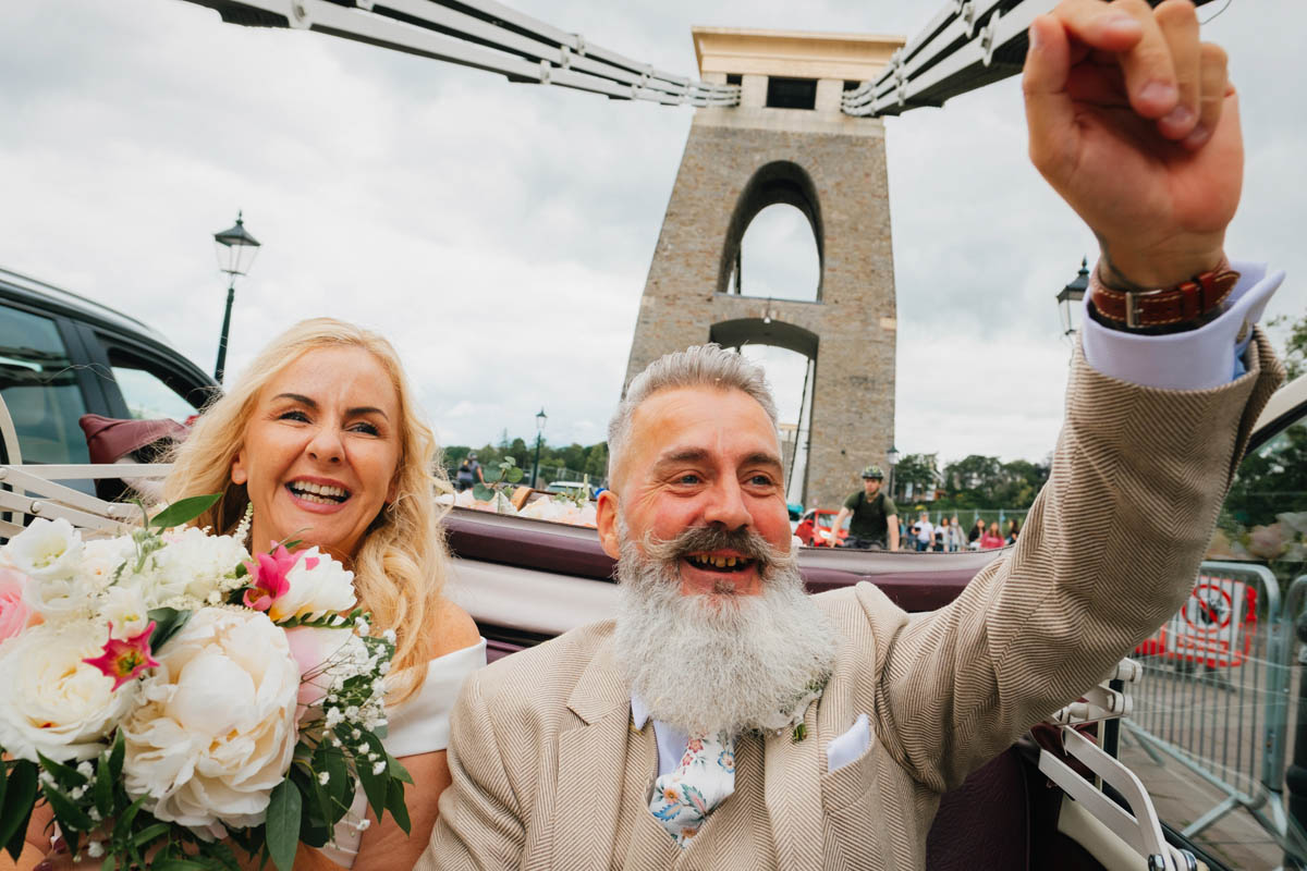 bride and groom wave and cheer whilst driving over the Clifton suspension bridge in a soft top car