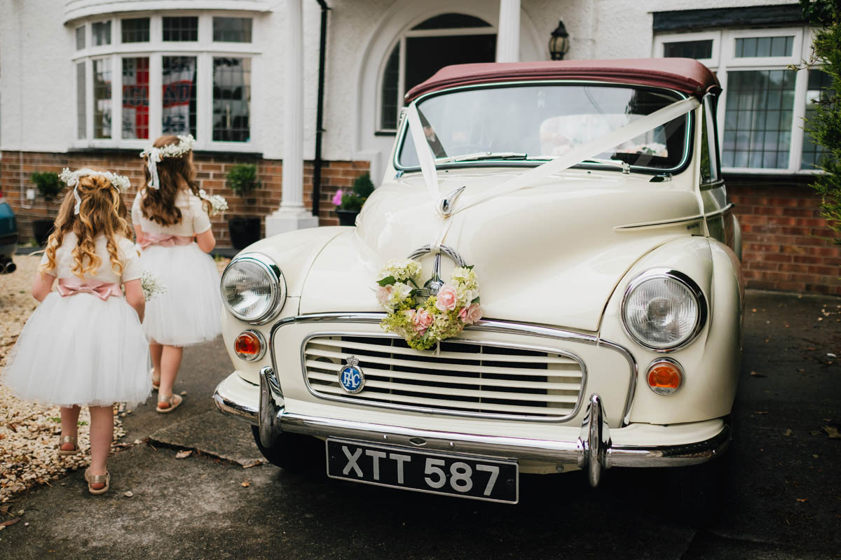 flower girls hold their bouquets in front of a Morris Minor adorned with wedding flowers