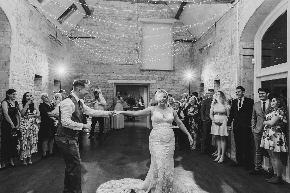 groom spins his new wife around the dance floor in black and white while wedding guests smile and cheer behind