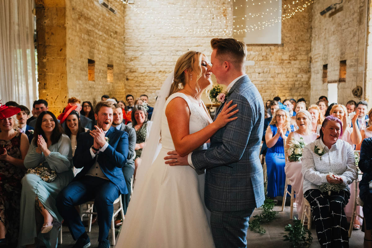 the newly-weds kiss as guest cheer behind in the ceremony room at Lapstone barn