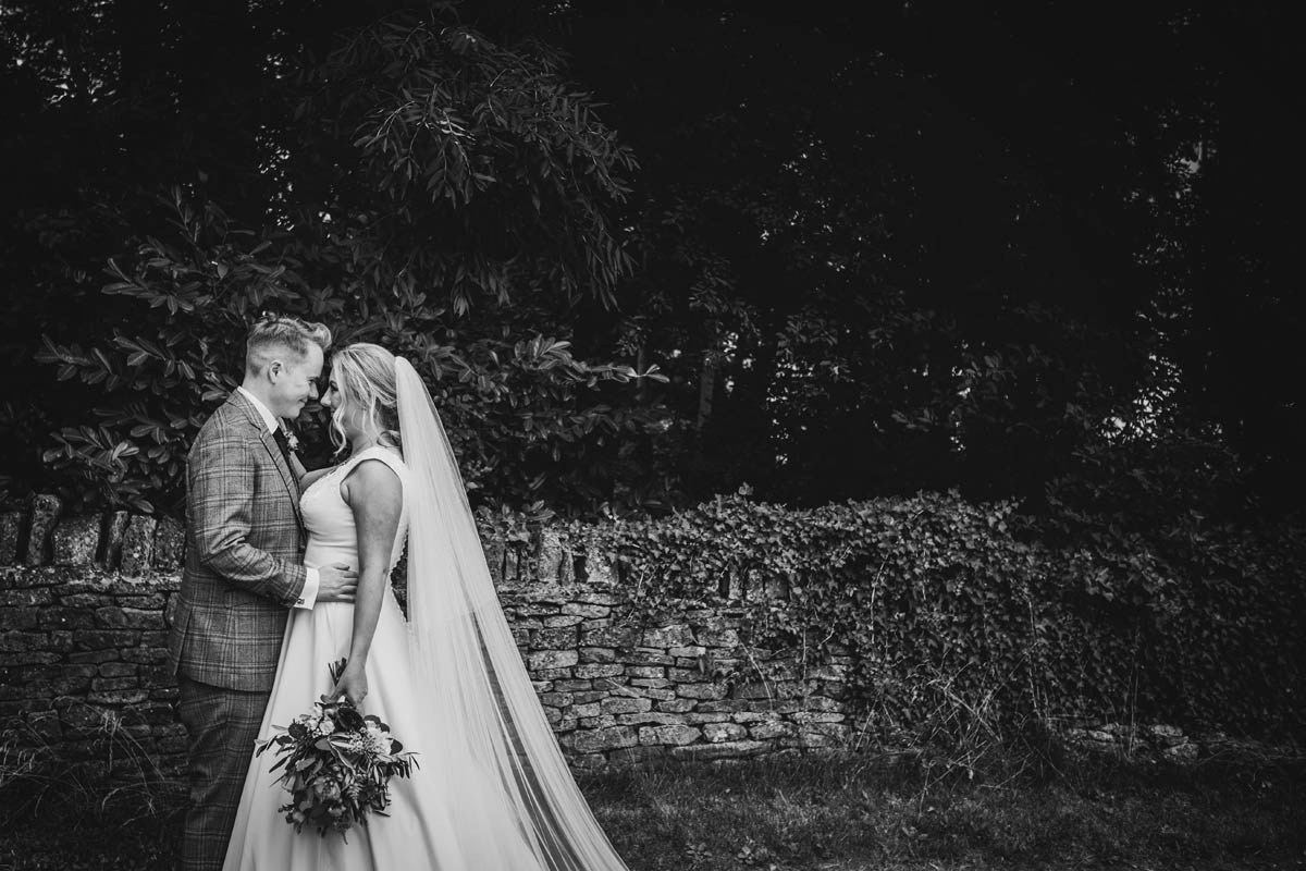 a romantic black and white photograph of the newly-weds looking into each other eyes in front of a stone wall and foliage