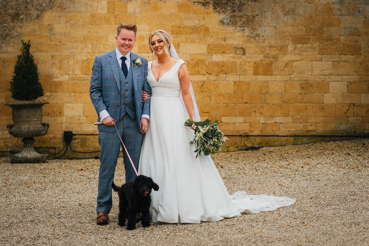the new husband and wife pose in front of a cotswolds wall with their cockerpoo dog