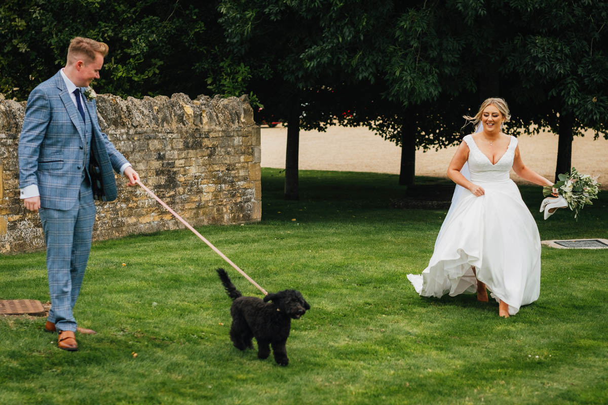 the bride runs after her new husband who walks their cockerpoo dog on a lead