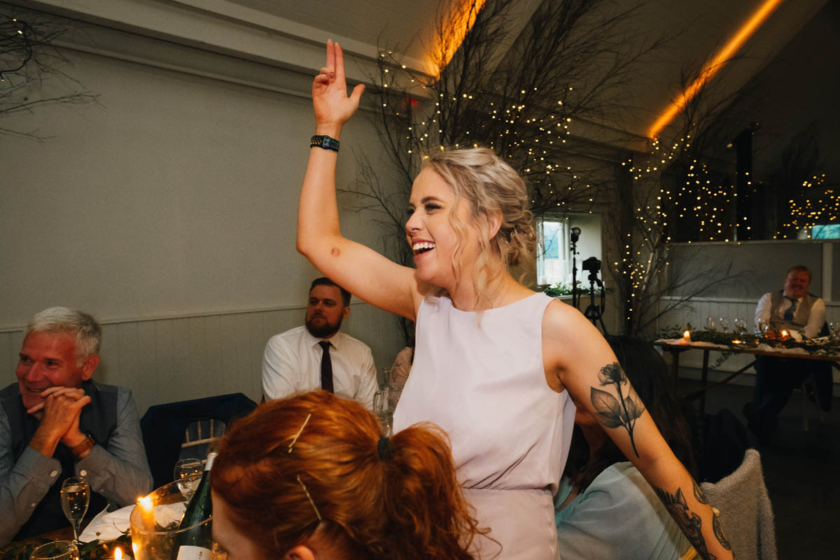 the groom's sister stands up front he table and waves her arms in the air as she laughs