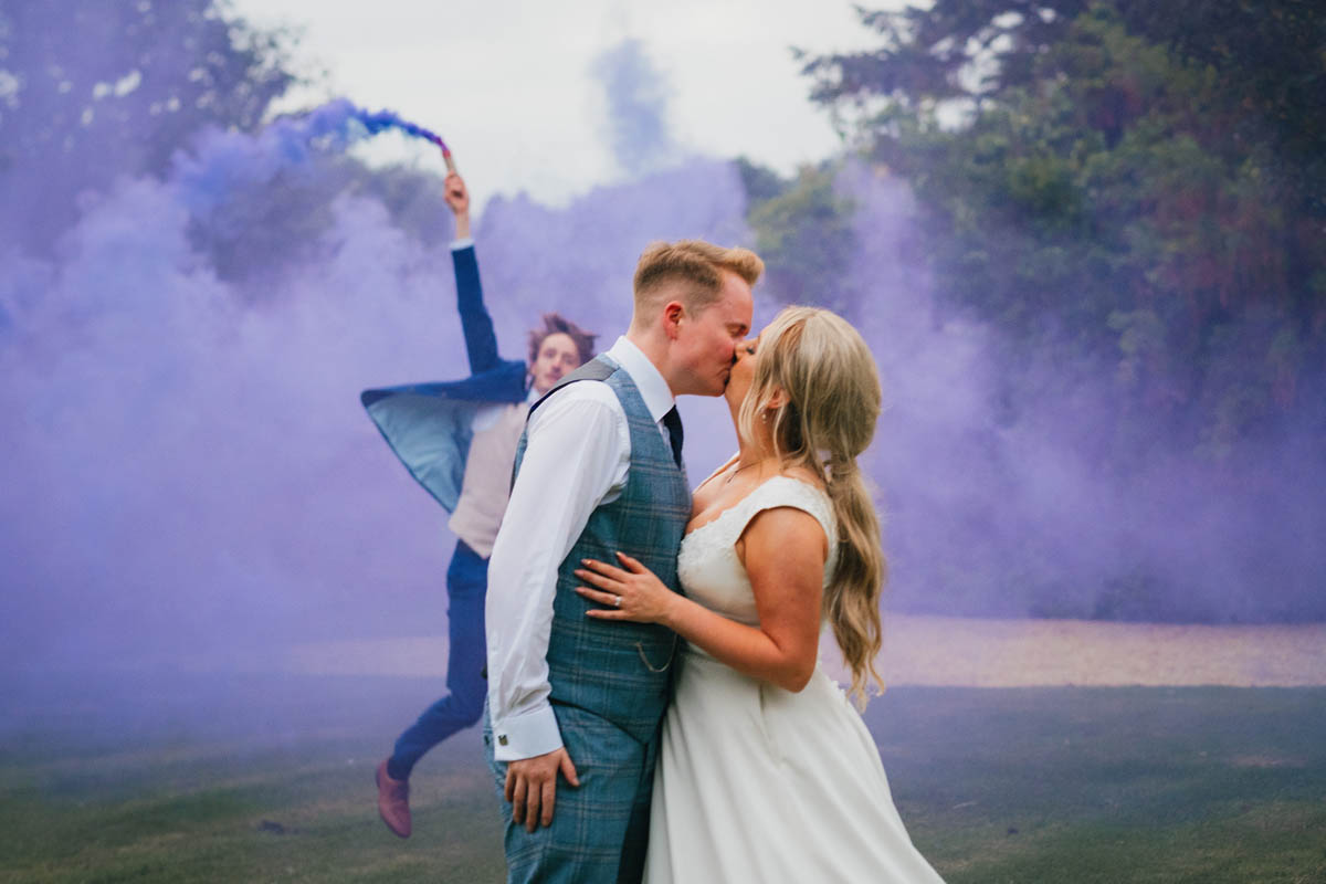 a groomsmen waves a smoke bomb behind the kissing bride and groom