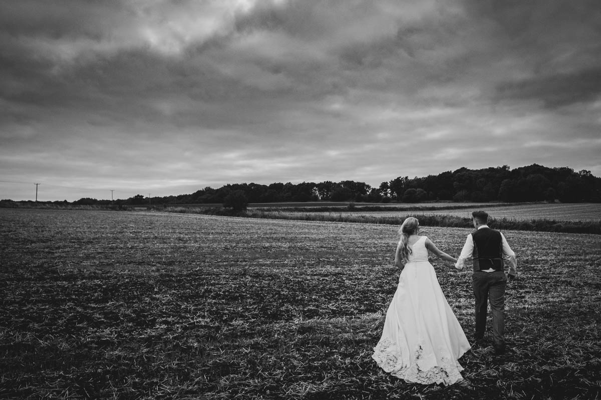 bride and groom walk away from the camera in a cotswolds field, the bride's gown trails behind