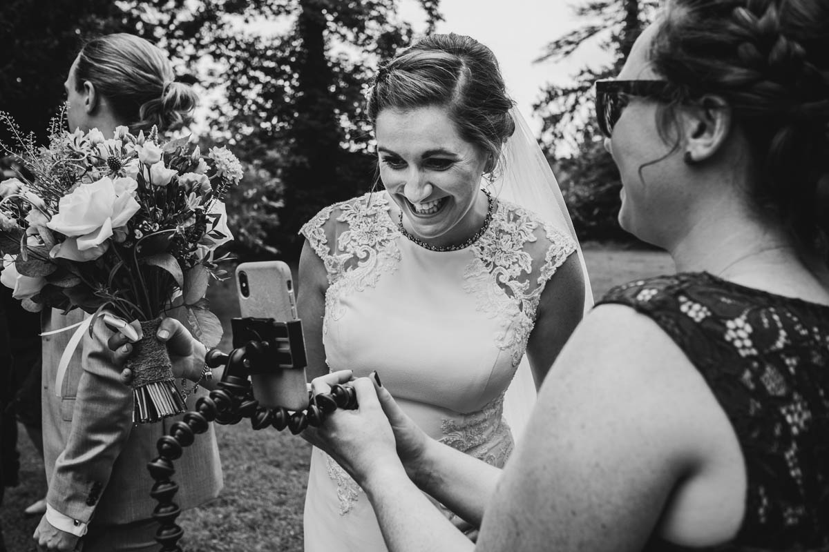a black and white photograph of the bride smiling at a phone as she chats to her brother via zoom on her wedding day