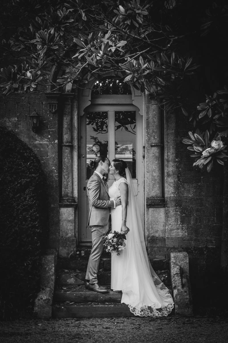 a black and white photograph of a newly-married couple kissing on the steps at Orchardleigh. Foliage frames them