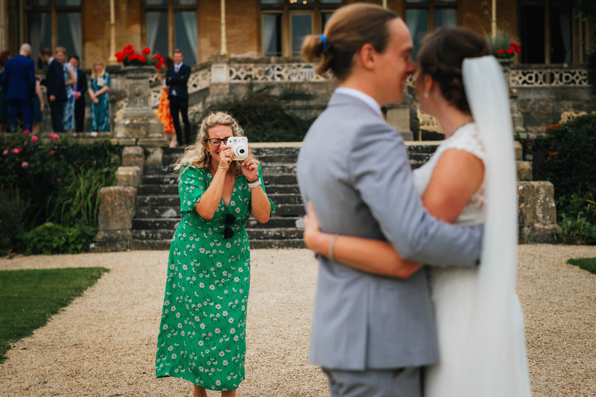 a guest uses a polaroid camera to take a photo of the bride and groom, wedding guests watch from the background