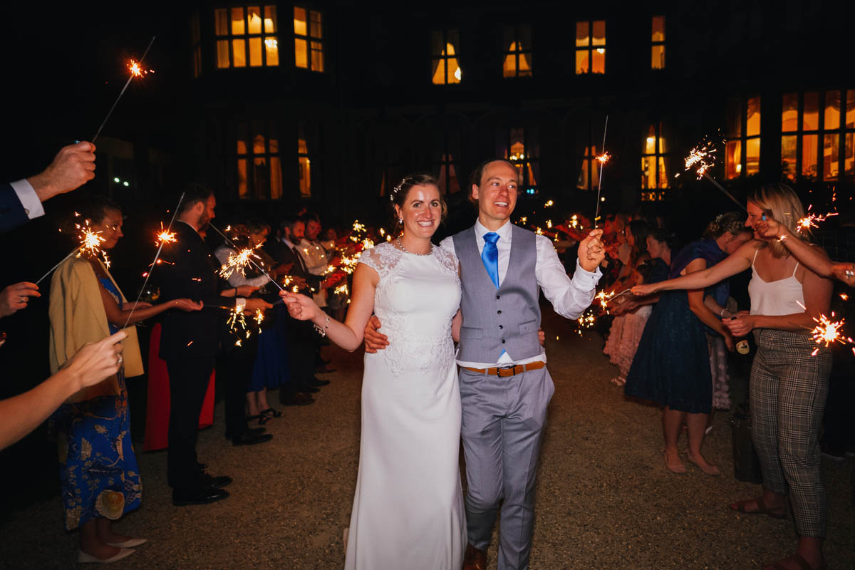 bride and groom walk down an aisle of wedding guests holding sparklers during their sparkler exit. The wedding venue fills the background