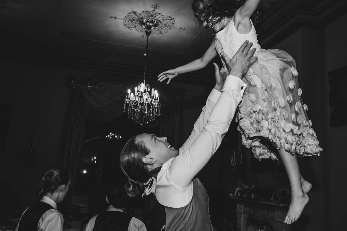 the groom throws a flower girl up into the air on the dance floor. A chandelier fills the background