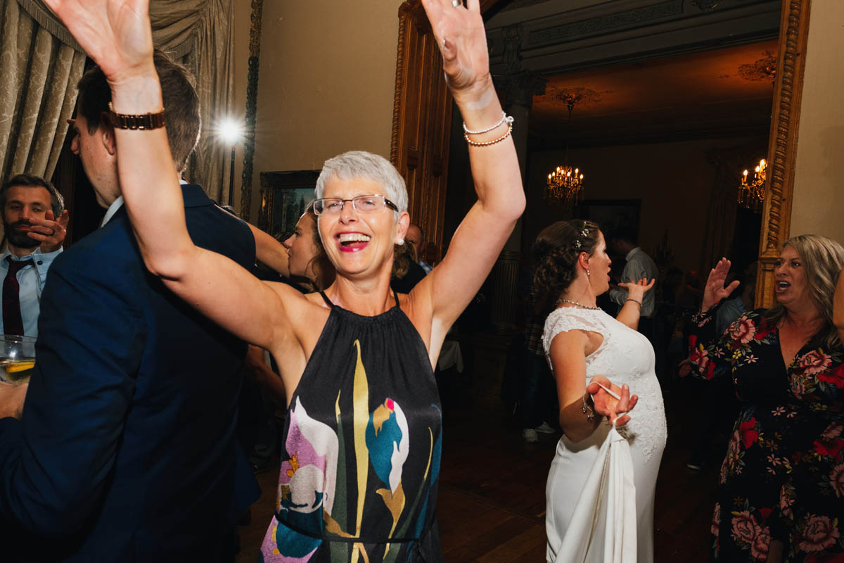 a woman smiles at the camera as they throws her arms in the air, a flash fires from behind and the bride and a wedding guest dance in the background