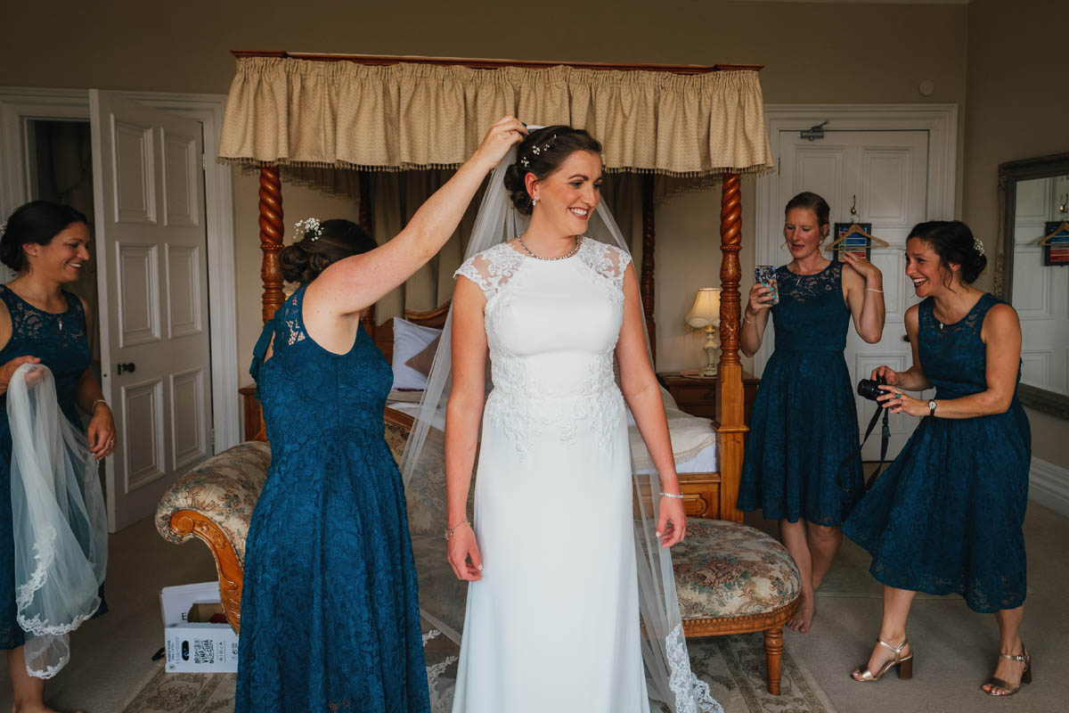 the bride stands in the middle of the room as bridesmaids take photographs whilst the maid of honour puts the veil into the bride's hair