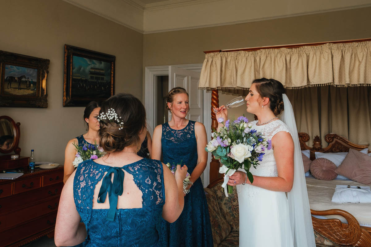 bridesmaids surround the bride as she downs the last of her champagne in the bridal suite