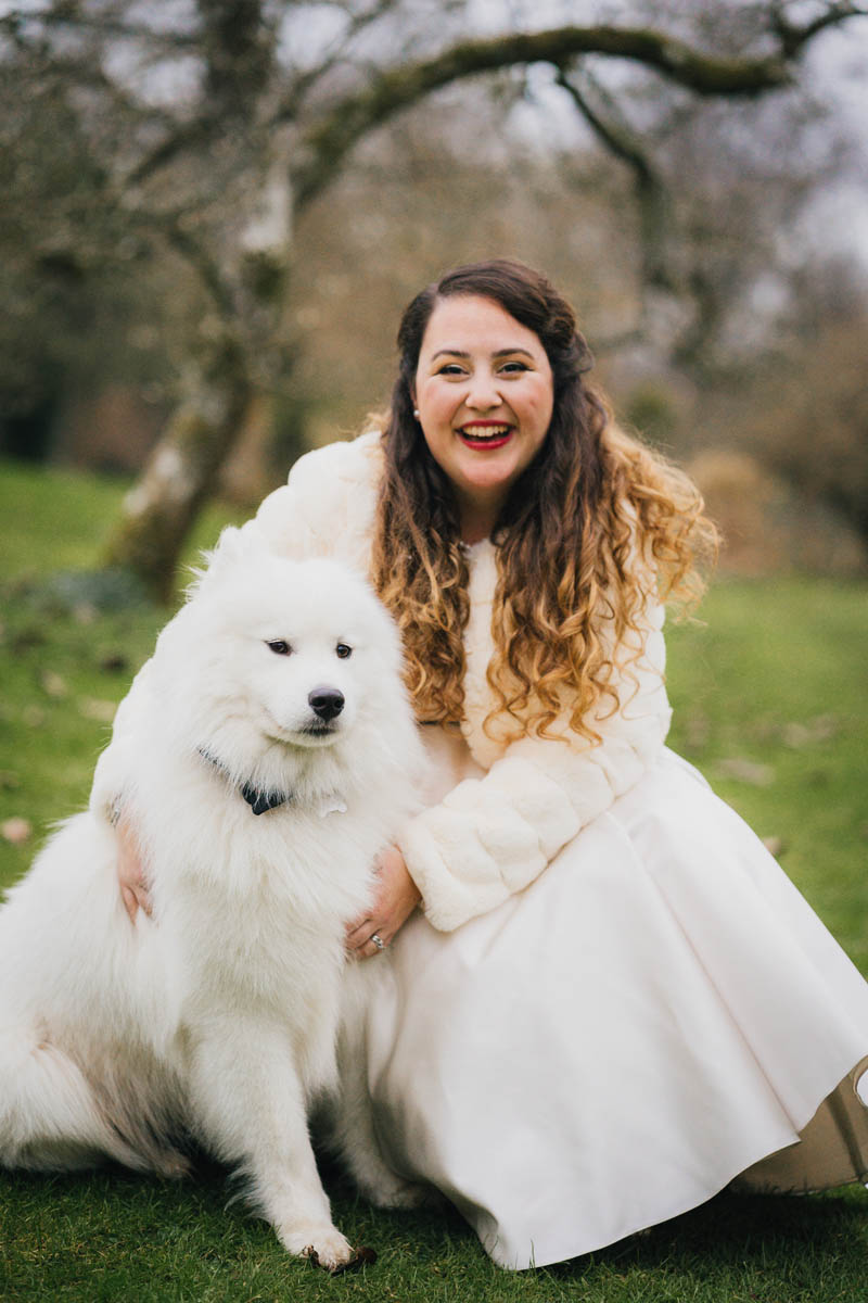 the bride poses with her white fluffy dog in a portrait picture, there is grass and a tree behind