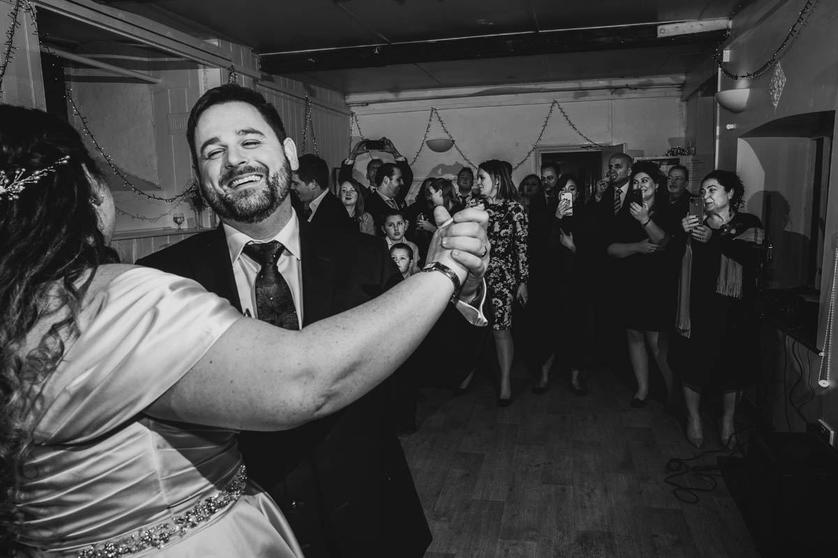 in the foreground the groom spins his wife around whilst wedding guests look on from behind, many of them record the dance on their phones