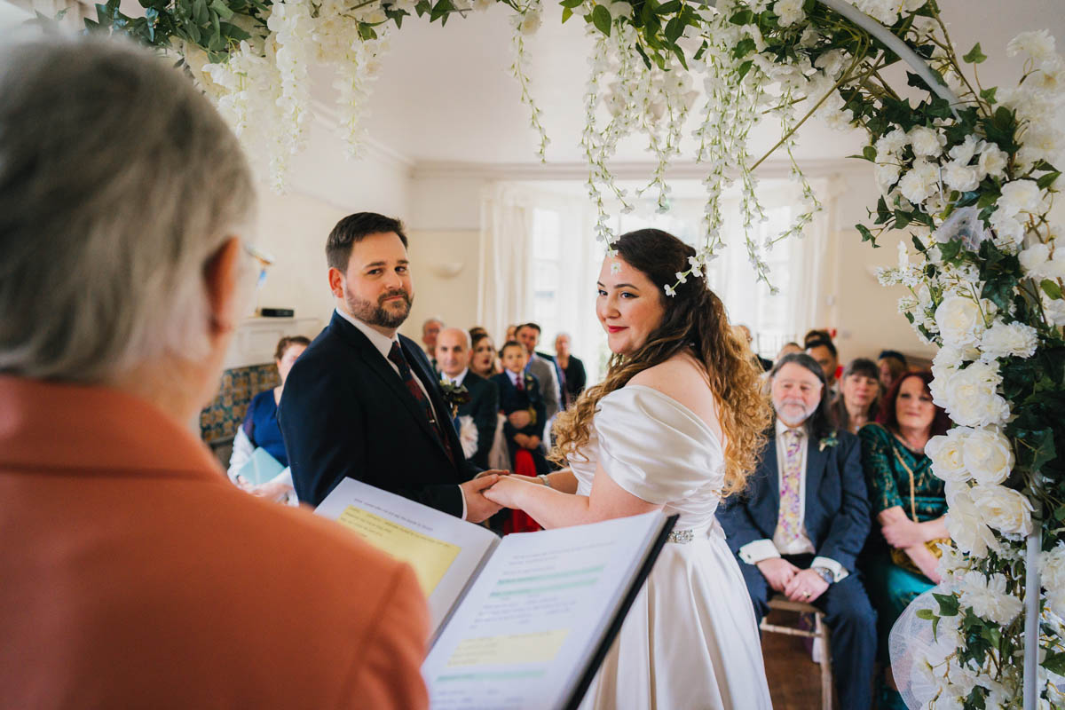 the marriage officiant holds her ceremony book in front of the man and woman getting married, they hold hands and look at the marriage officiant. wedding guests watch being