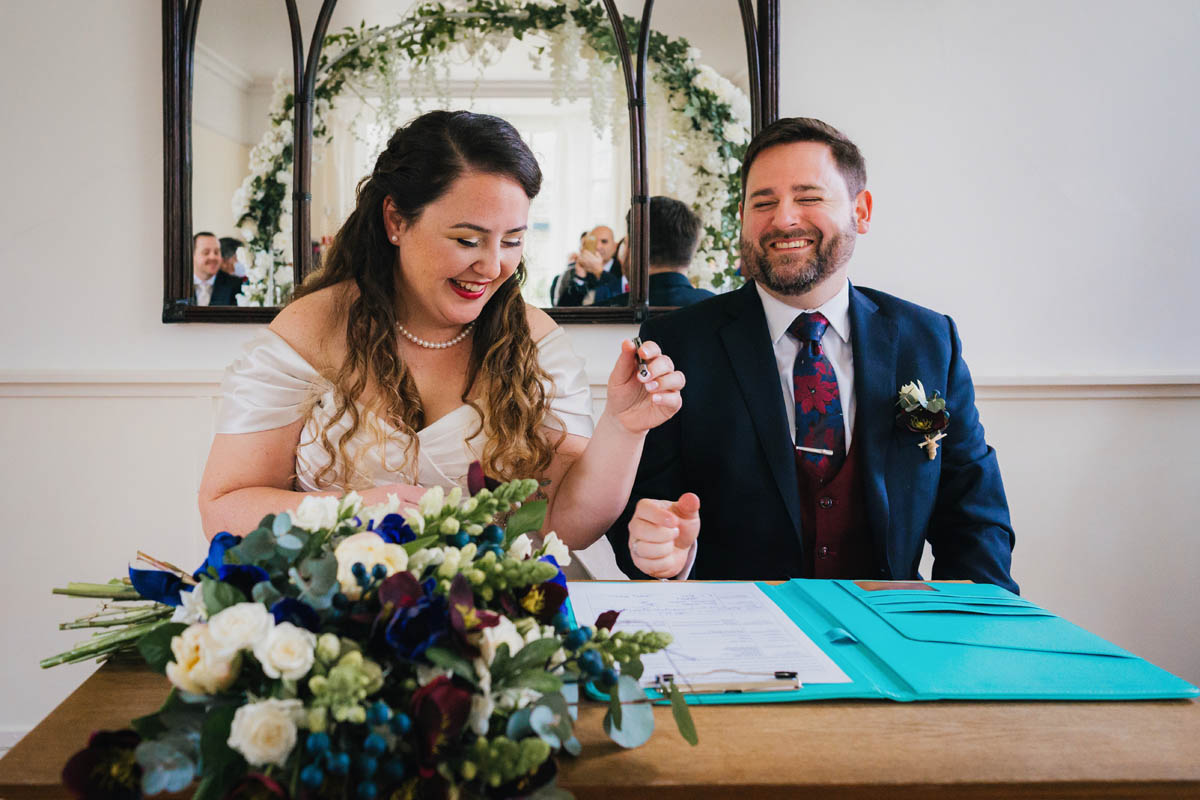 the bride holds the pen above the marriage certificate while the groom laughs. a mirror is behind and shows smiling guests./ the bouquet sits on the table