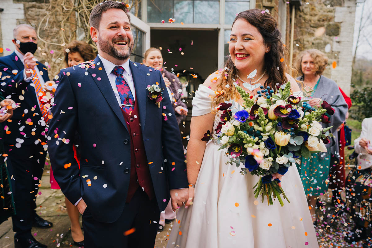 bride and groom holds hands and laugh as guests smile and throw confetti over them
