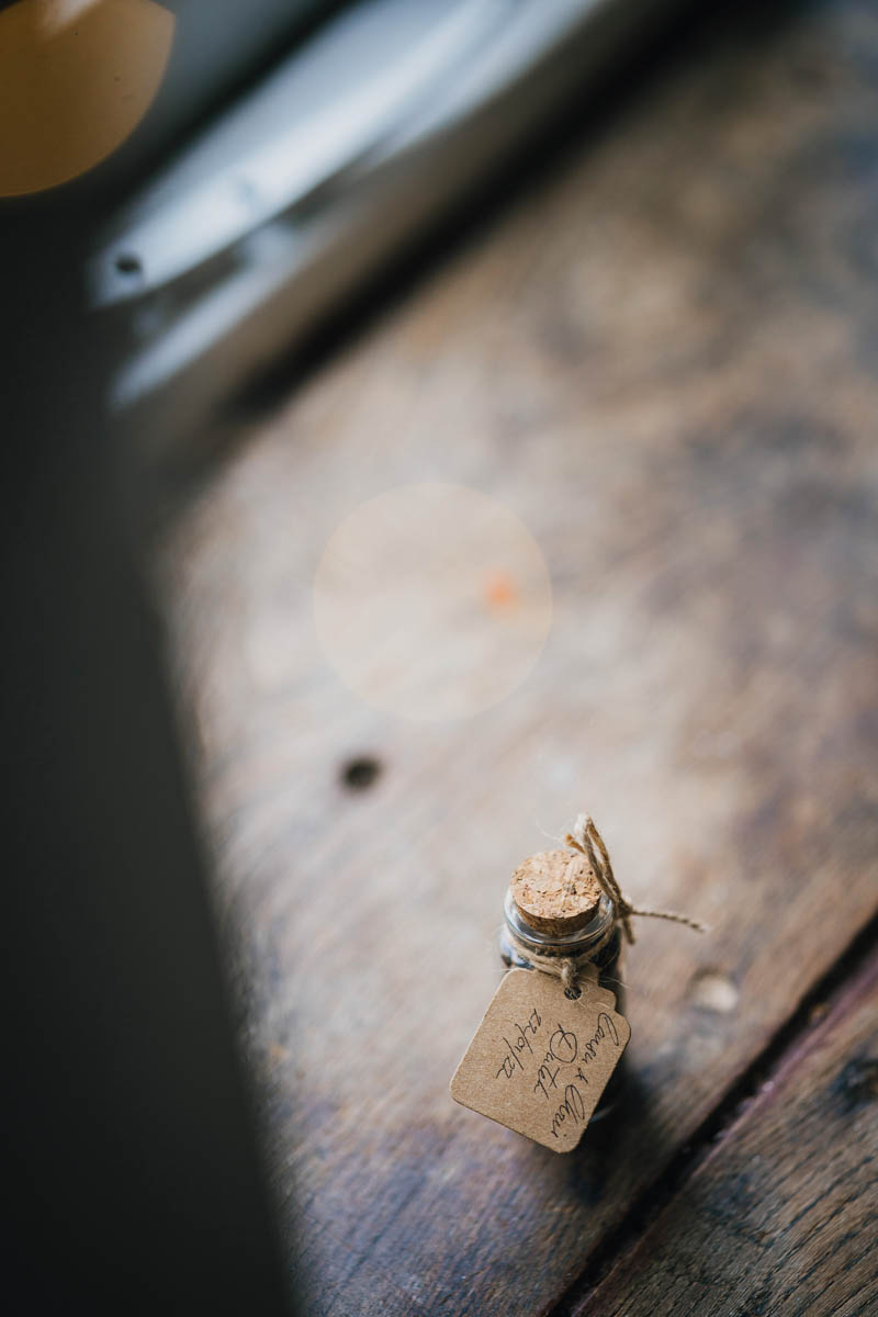 a wedding favour of a corked jar with brown string and a craft paper label sits on a wooden window sill