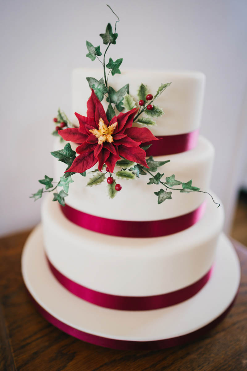 a three tiered wedding cake with red ribbon around each tier, a beautiful sugar craft flower and ivy sit between the top two tiers