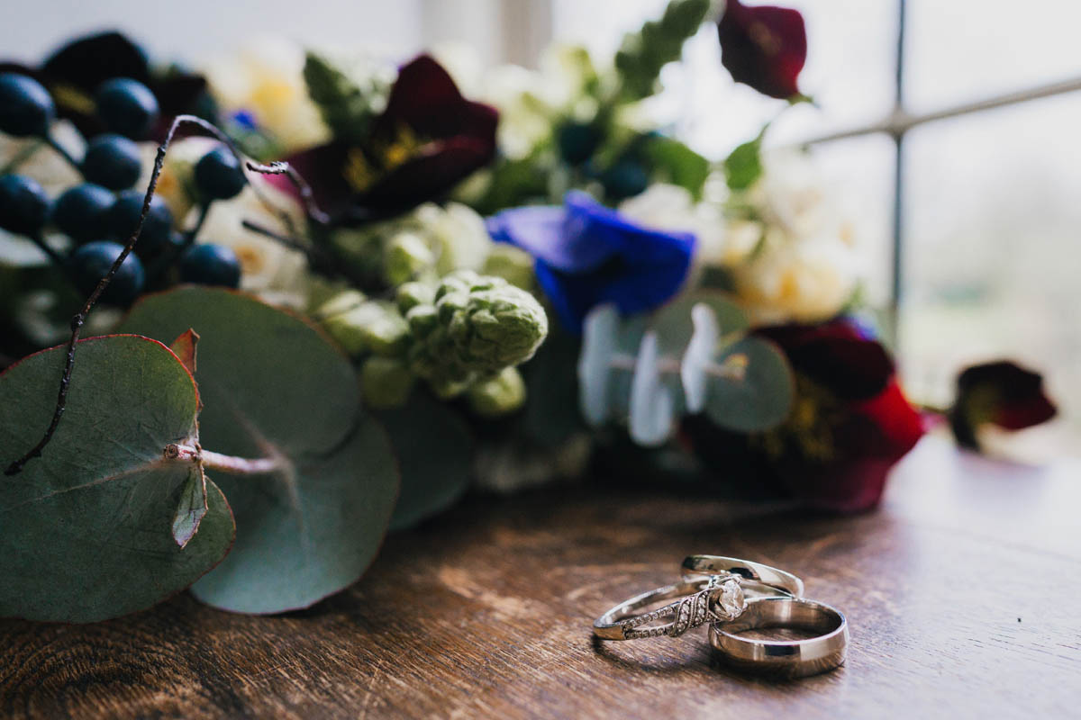 close-up photograph of the two wedding rings and engagement ring, the bride's bouquet and a window fill the background
