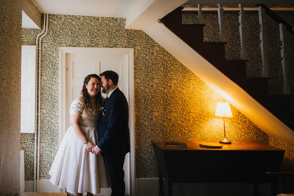 bride and groom hold hands under the stairs, they are hit with dingo light from the left and light from a lamp from the right, a set of stairs lead upwards on the right