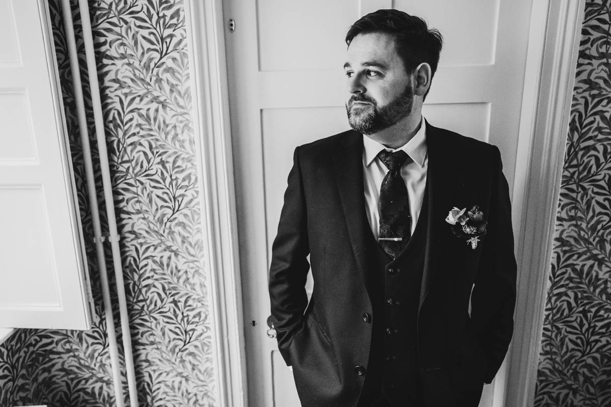 black and whit photograph of the groom with his hands in his suit pockets, he gazes out of the window to his side. he stands in front of a white wooden door with wallpaper covered in foliage