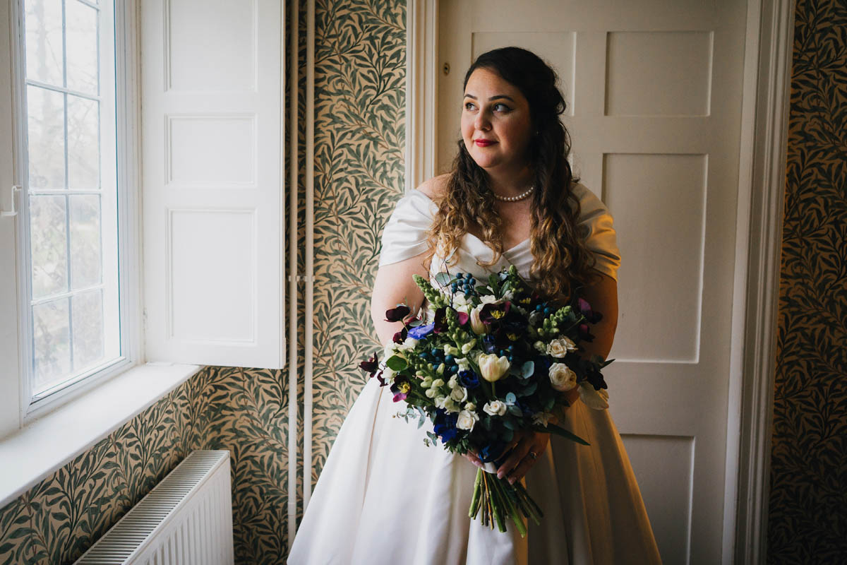 the bride stands in a window which she peers out of whilst holding her bouquet, the light hits the side of her face. behind her is a white wooden door and a wallpaper covered in greenery