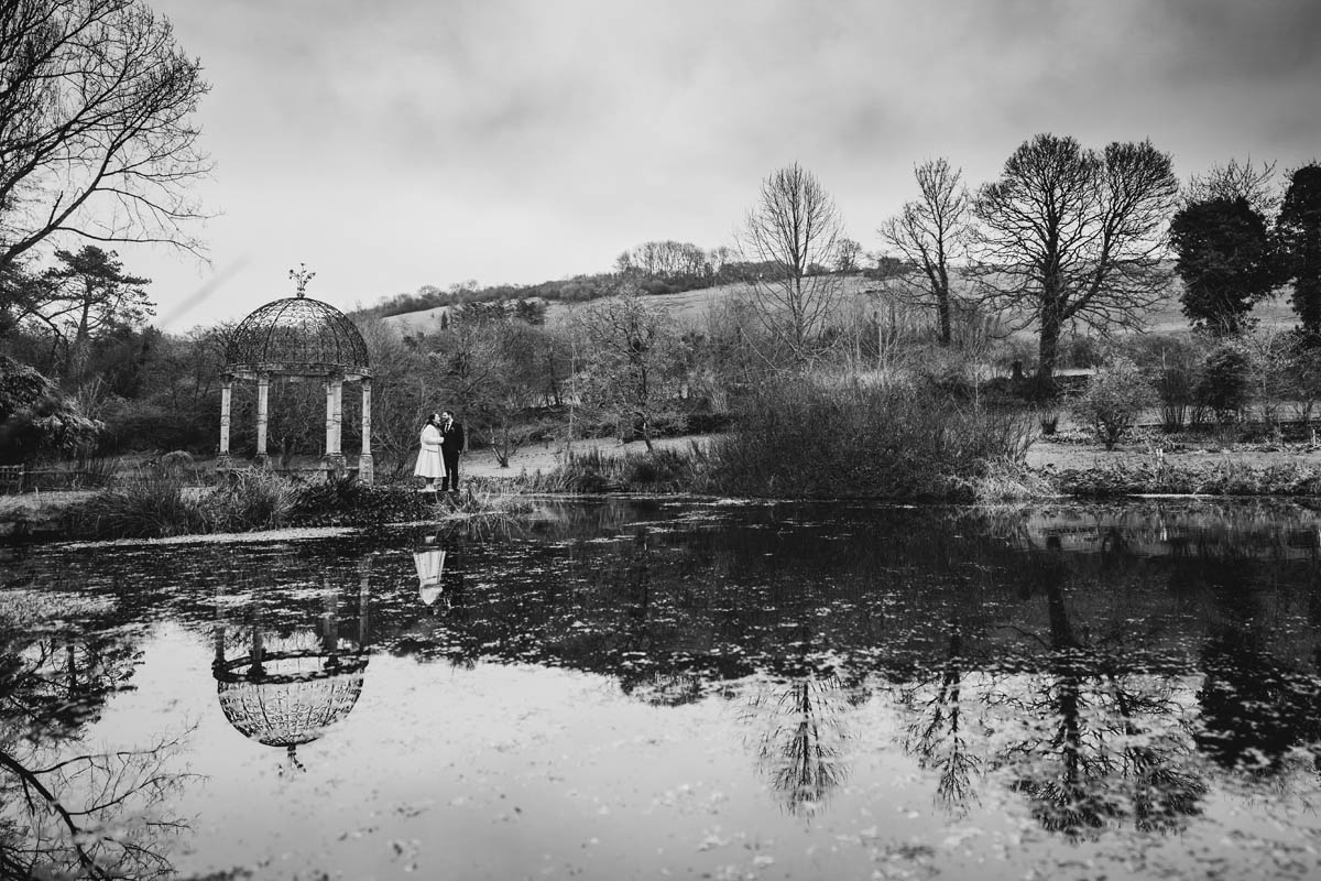wide angle shot of the lake at springhead trust. The bride and groom share a kiss next to the lake, the reflection appears in the clear water below them