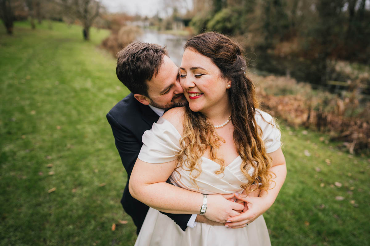 the groom hugs his bride from behind and nuzzles into her neck, she rests her cheek on his and smiles