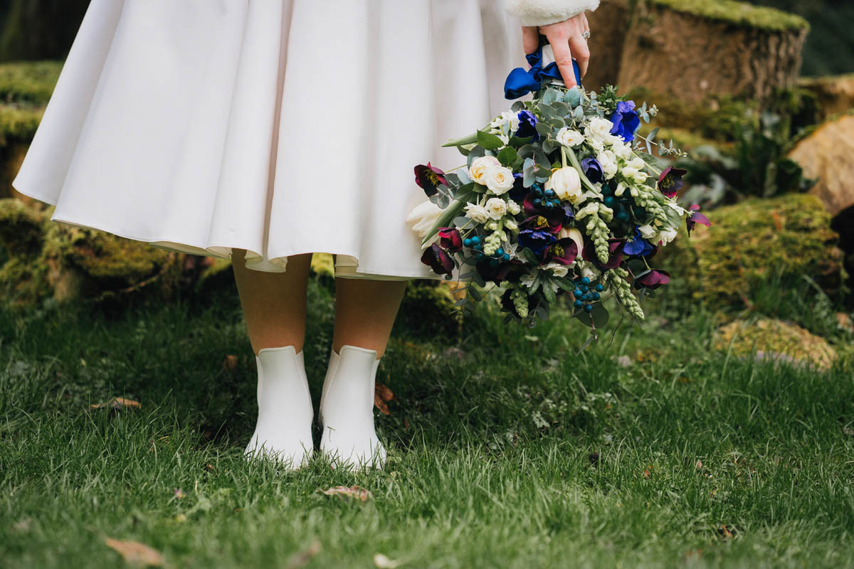 a close up photograph of the bottom of the bride's wedding dress, her wellies and bouquet, in front of a pile of mossy logs
