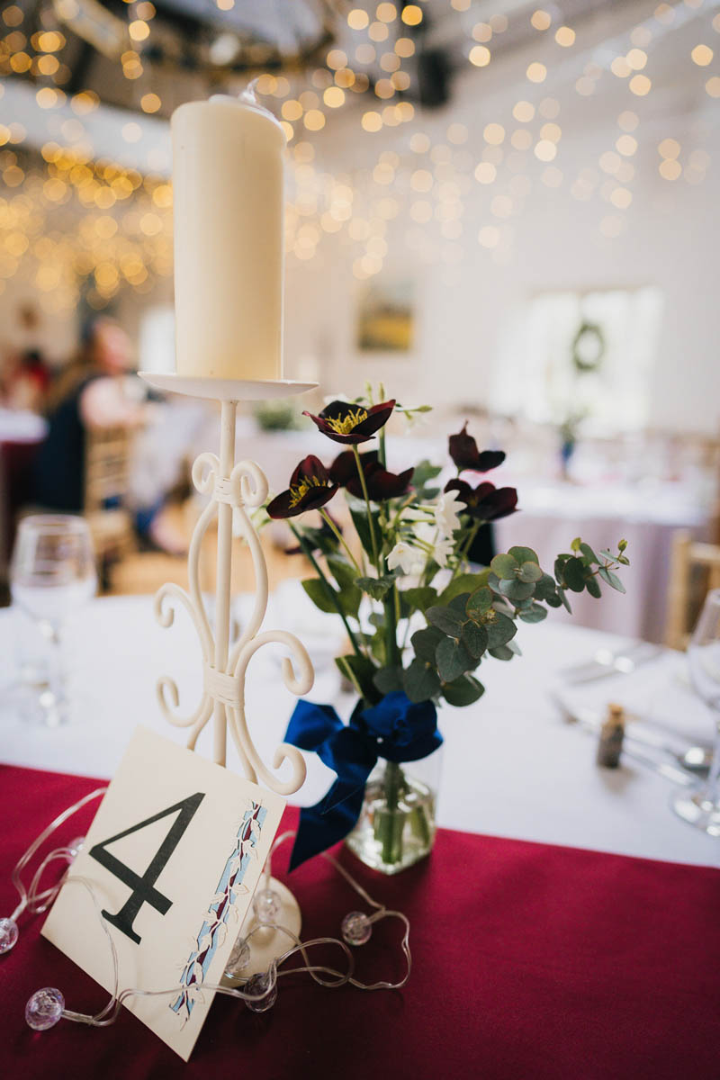 a candle, flowers in a vase with a blue ribbon, a table number and fairly lights are the centre pieces of the tables for the wedding breakfast