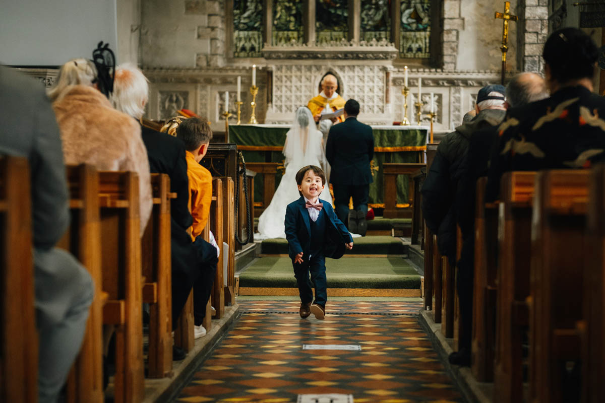 the bride and groom's son runs down the church aisle while the bride and groom are blessed by the vicar in the background