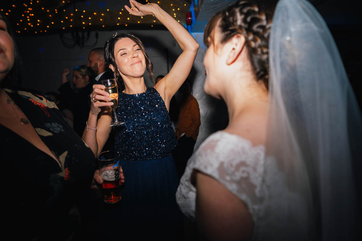 a bridesmaid throws her arms in the air as she dances with the bride. Fairy lights create bokeh behind