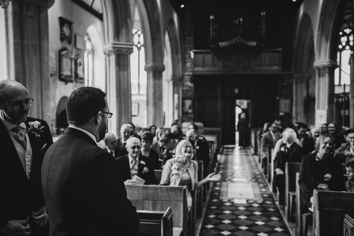 the groom and his best man stand in the foreground. in the background wedding guests sit in pews and await the bride's arrival