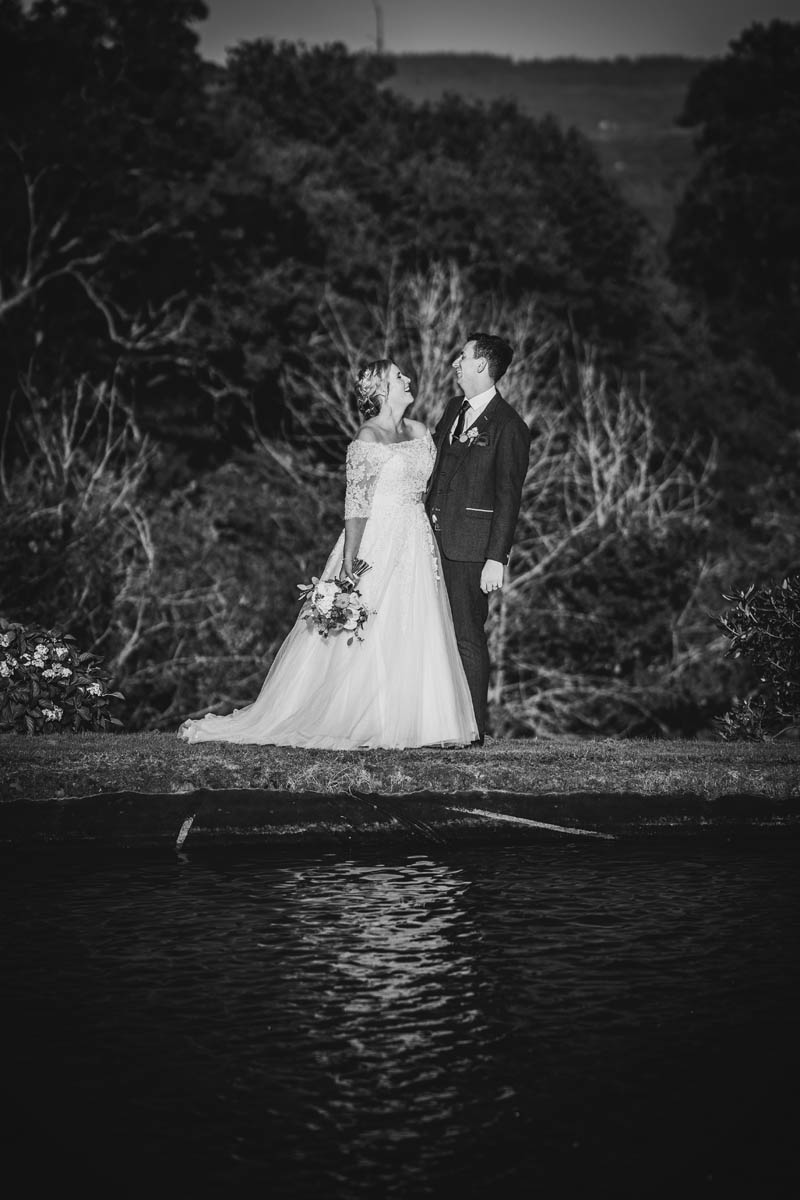 the bride and groom throw their heads back in laughter in front of the lake at Upton Barn, the backdown hills are in the background. a black and white photograph.