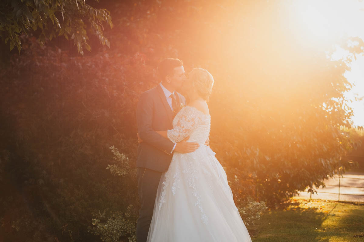 sun flare hits the newly-married couple as they share a kiss in front of some bushes