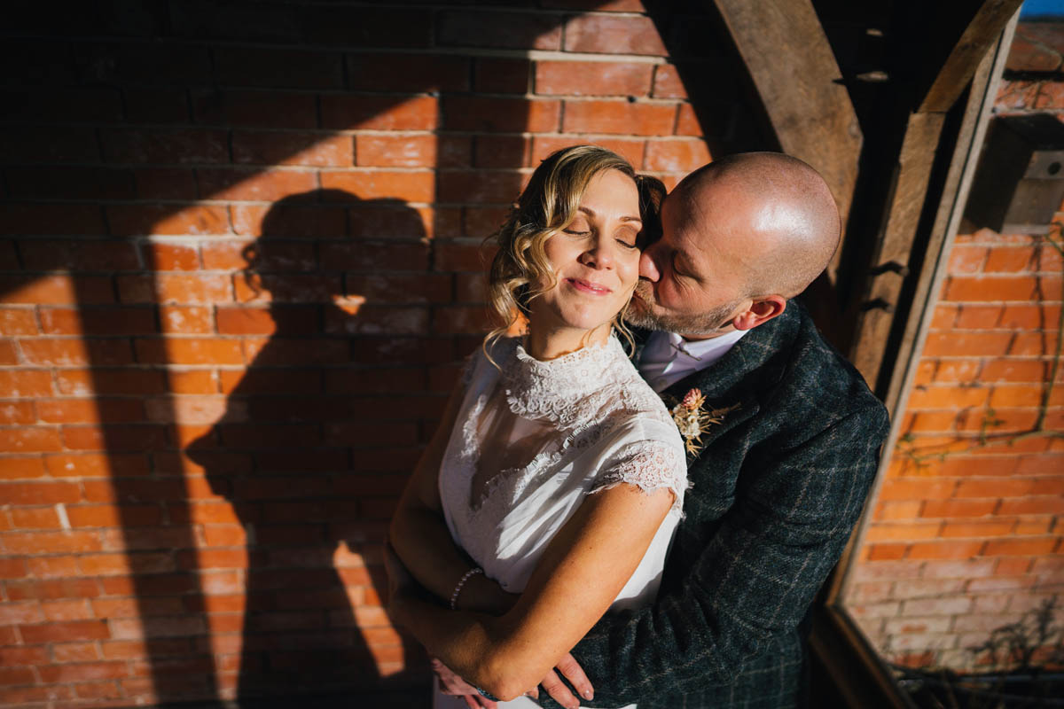 light hits the groom as he hugs his new wife from behind. She rests her head on his cheek as he kisses her. Their figures cast a shadow on the redbrick wall behind an they are framed by oak timer