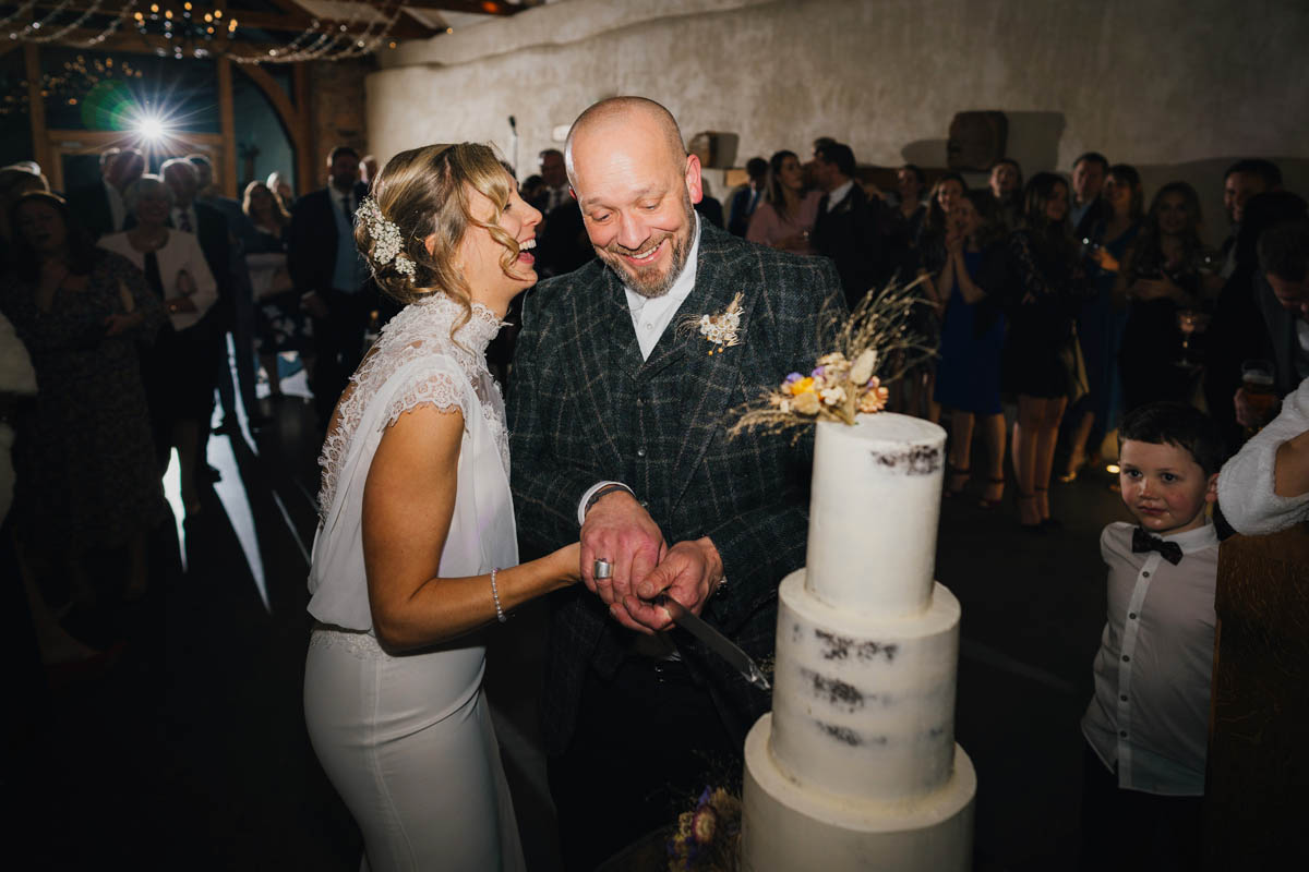 the bride and groom hold a knife together as they cut through their 3 tiered wedding cake, the bride laughs and her husband smiles