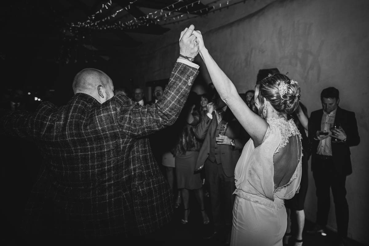black and white photograph of a bride and groom raising their hands in the air on the dance floor. Guests whistle and cheer behind