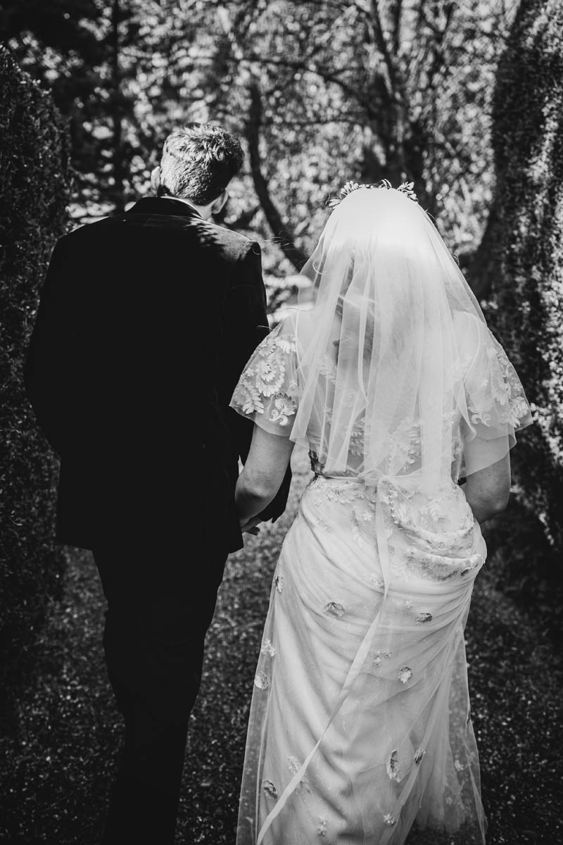 the newly-wedded couple walk up the tree-lined path towards their wedding venue. Shot from behind and in black and white