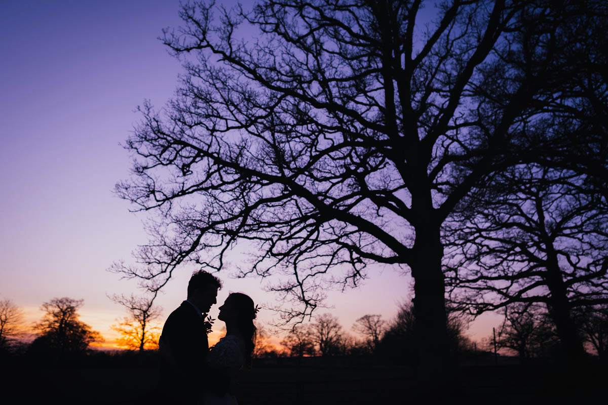silhouette of the newly-weds and a large tree. The sky is blue and purple as the sun sets
