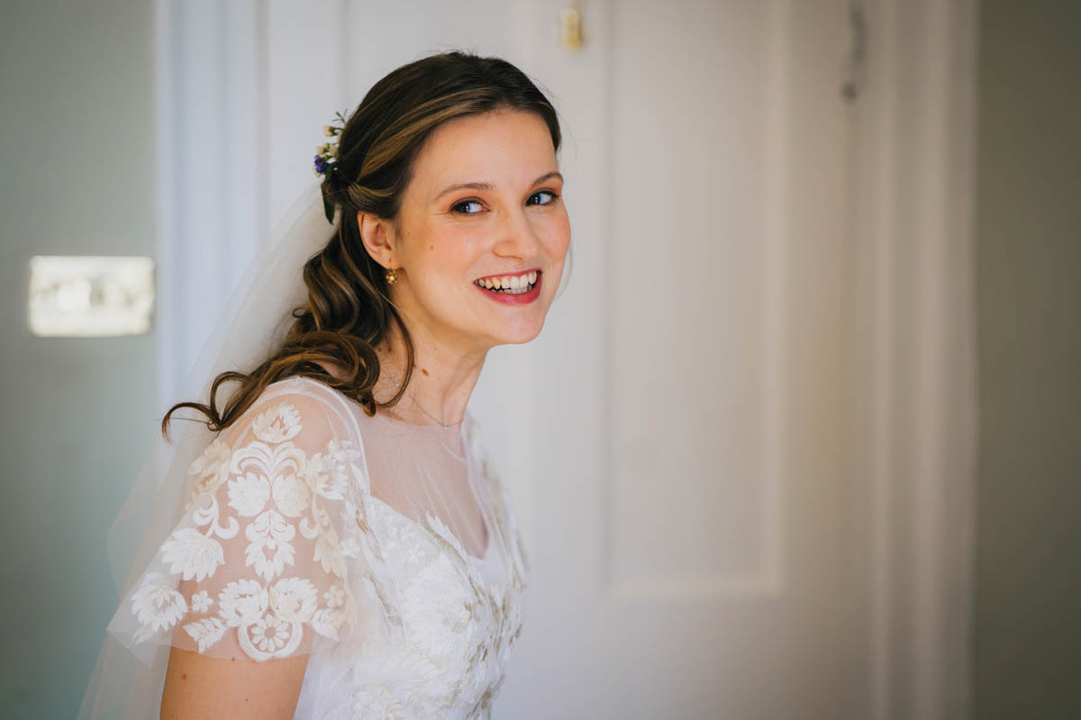 bride pulls a cheeky grin as she looks beyond the camera, she is dressed in a lacy capped sleeve wedding dress with veil and flowers in her curled hair