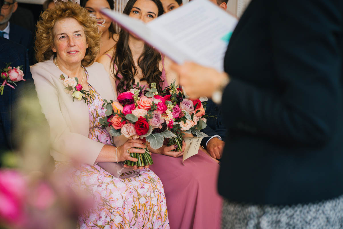 the bride's mother seated in the front row of the wedding ceremony. she holds the bride's bouquet as her daughter ties the knot
